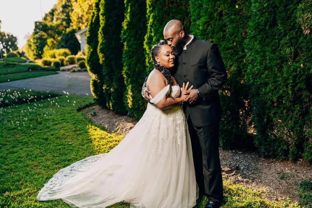 A couple embraces in a romantic garden setting with lush green trees and sunlit pathways.