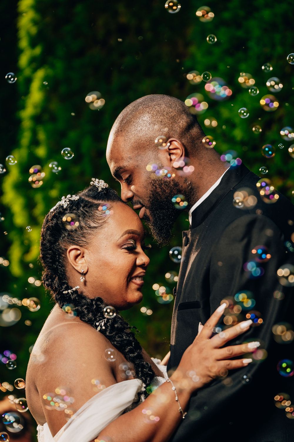 A romantic wedding couple shares an intimate moment surrounded by floating soap bubbles in a garden setting.