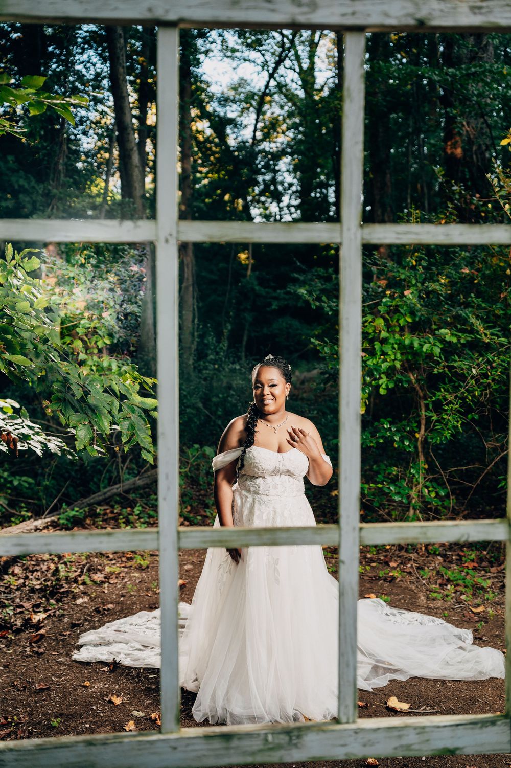 A bride in a white wedding dress sits gracefully on a stone ledge surrounded by lush greenery and window panes.