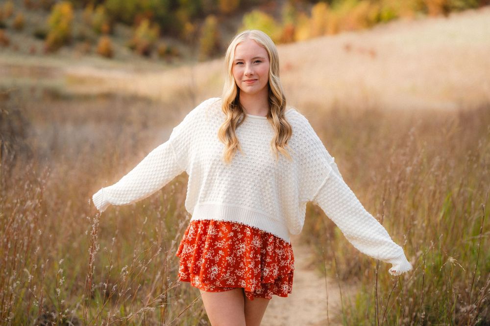 Young woman in white sweater and floral skirt poses with arms outstretched in autumn field at sunset.