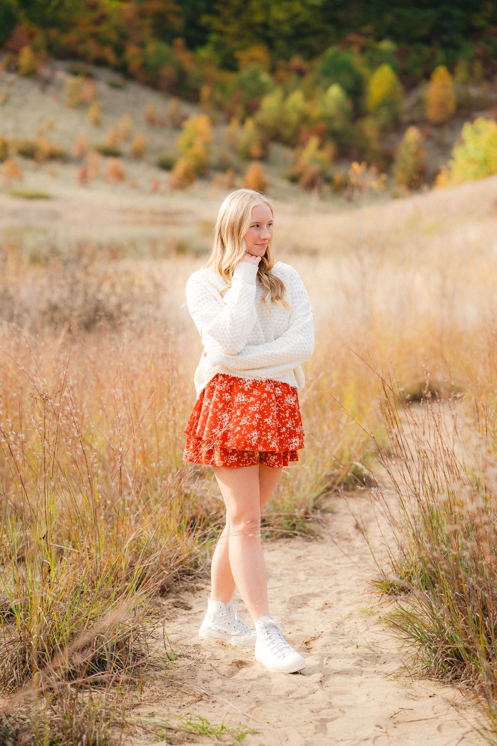 A person in a white sweater and red floral dress twirls playfully in a golden autumn field at sunset.