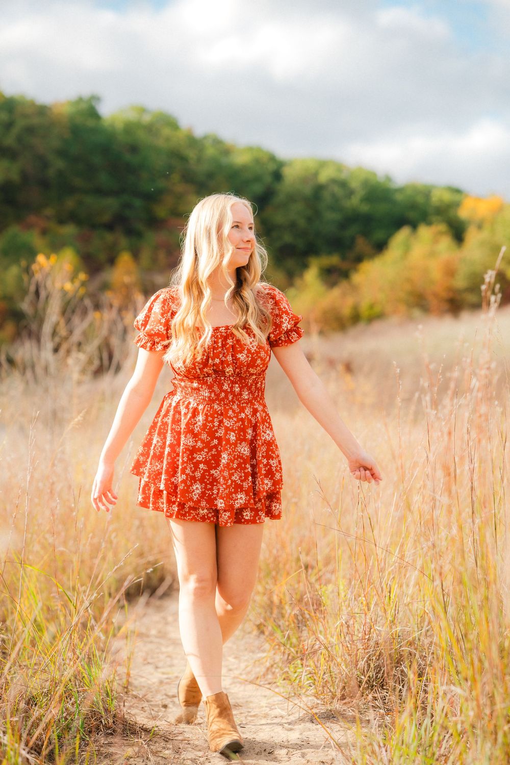 A woman in a red floral dress walks through a golden autumn field with tall grass and trees in the background.