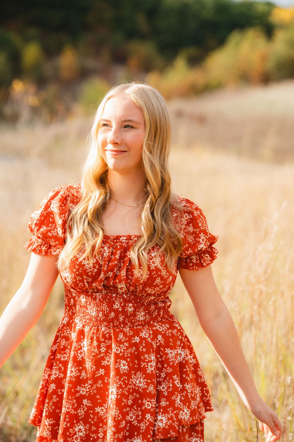 A woman wearing a red floral dress stands in a sunlit meadow surrounded by autumn foliage.