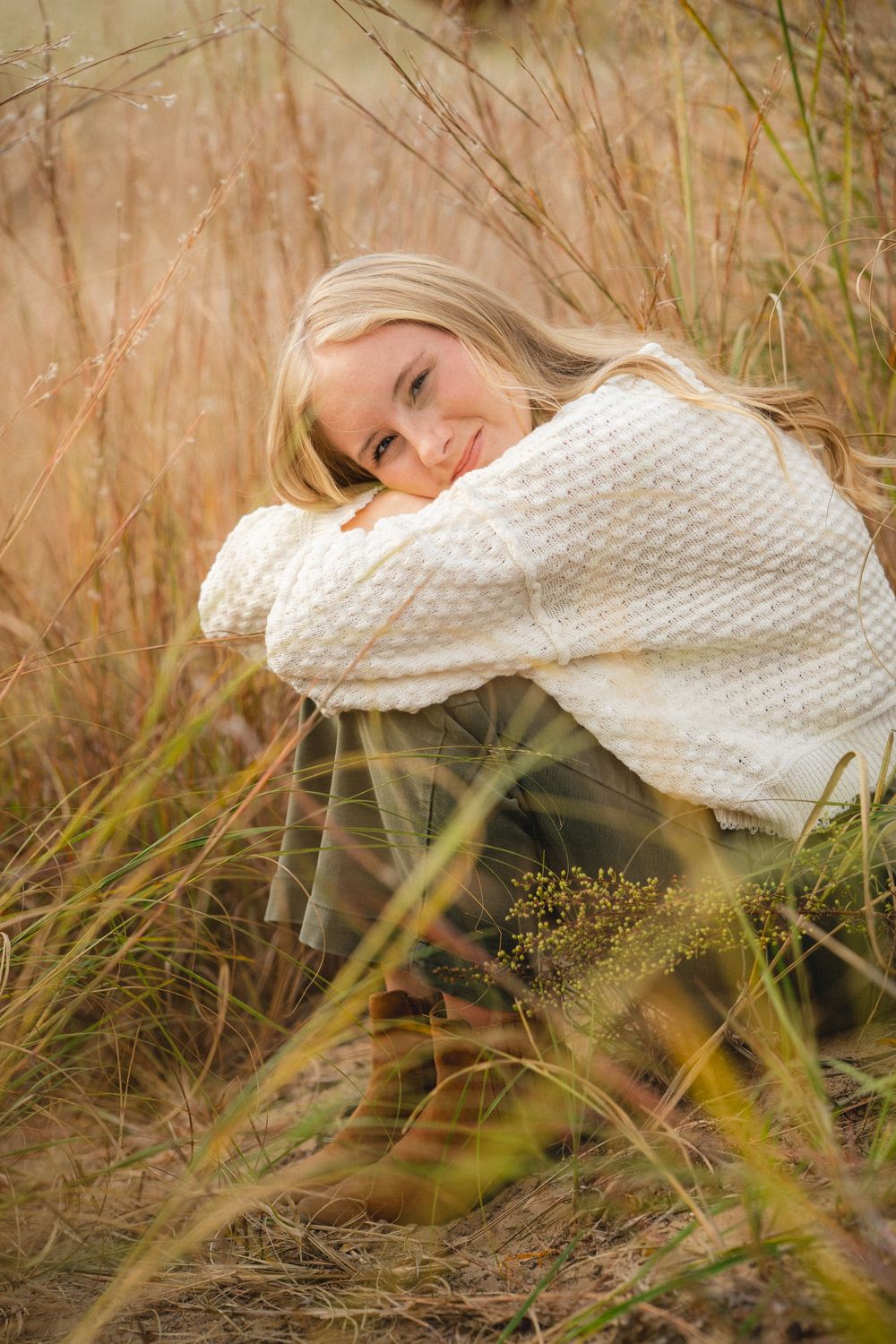 A woman in a white knit sweater rests among tall grass in a natural outdoor setting.