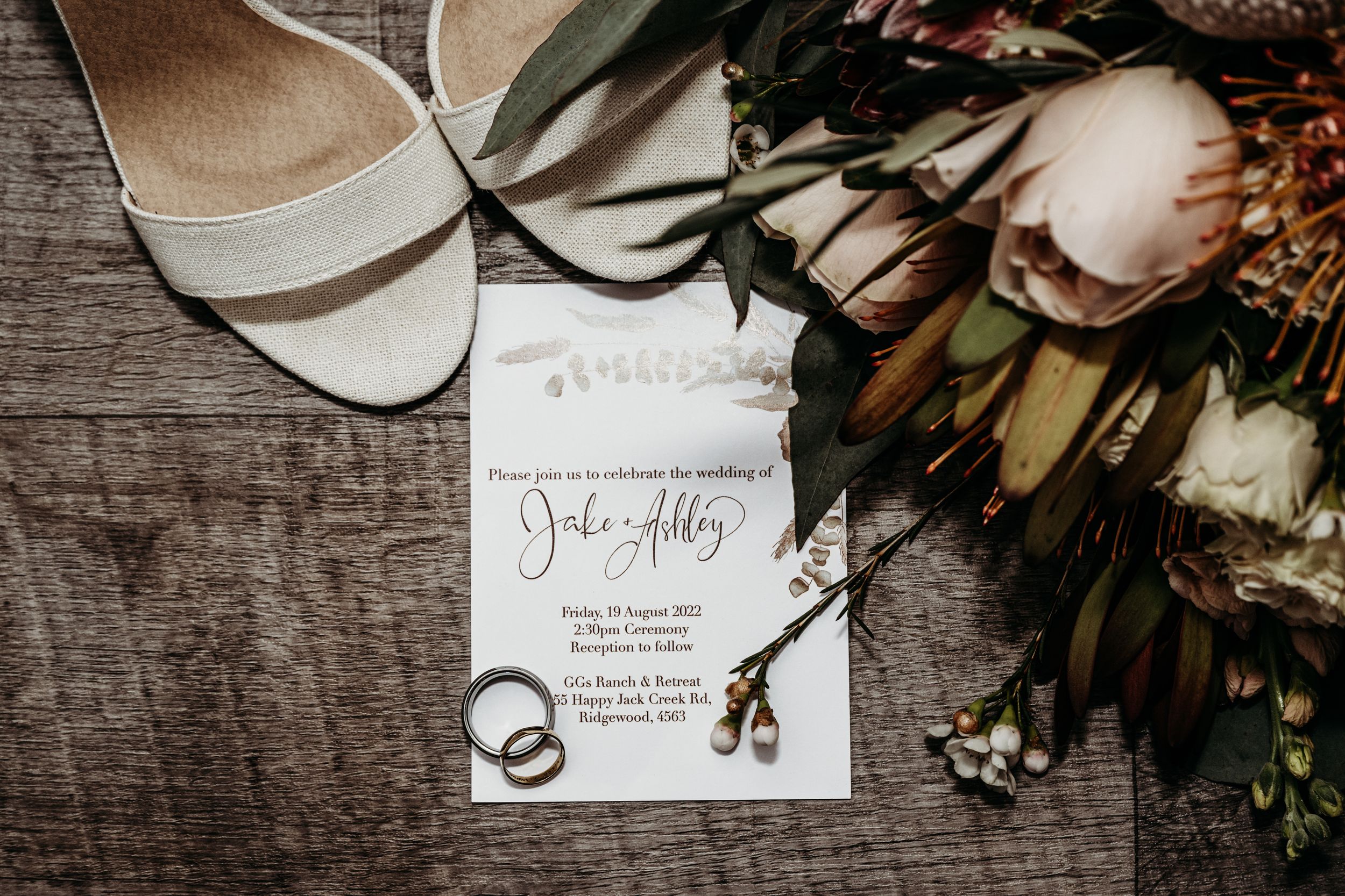 Wedding details including white bridal shoes, invitation card, wedding rings and floral bouquet on rustic wooden surface.