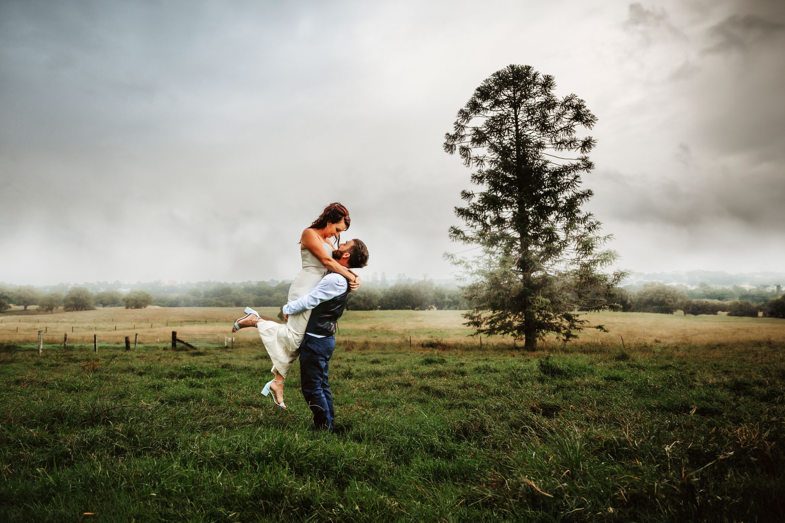 A couple embraces in a romantic pose on a grassy field with a lone tree in the background.