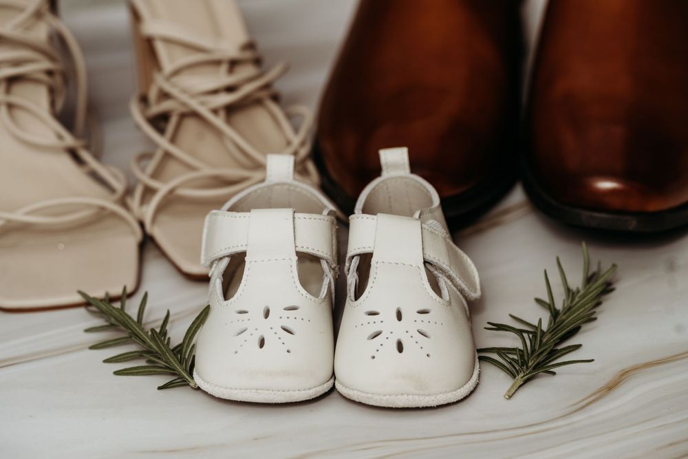 White baby shoes arranged with brown dress shoes and greenery on light background.