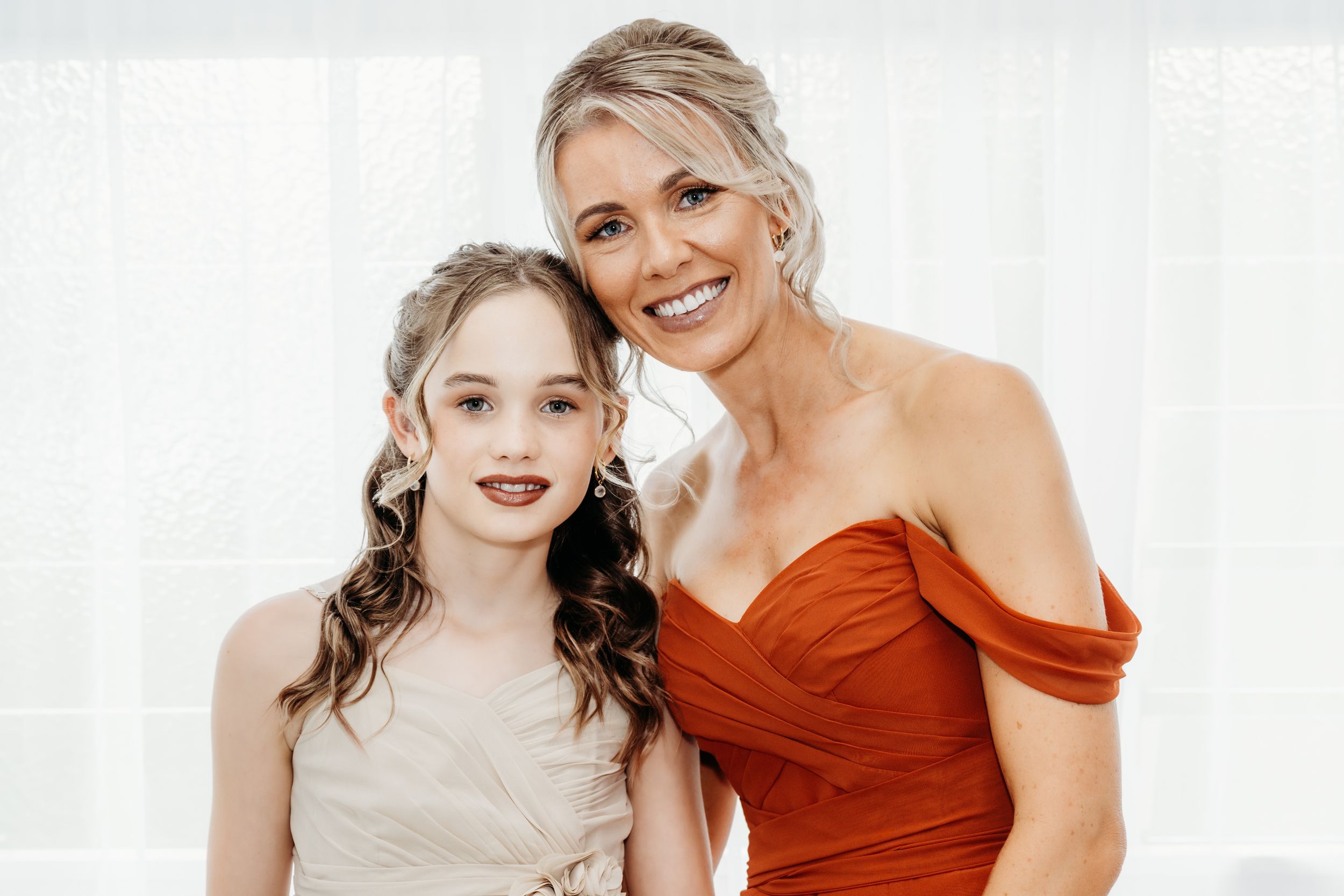 Two people in formal dresses, one wearing red and one in beige, pose together indoors.