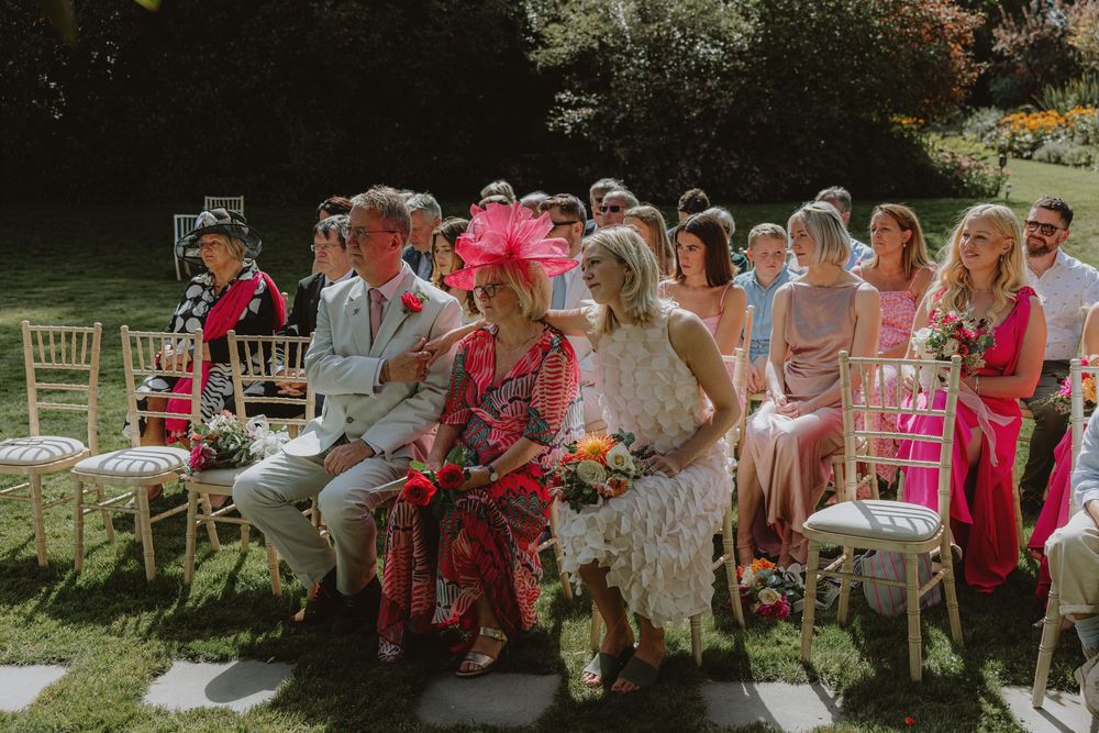 Wedding guests seated outdoors in pink and floral attire during ceremony.