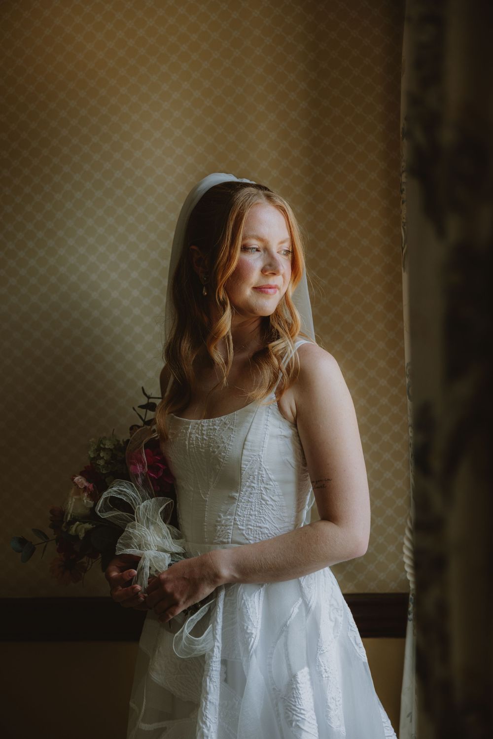Bride in white lace dress poses indoors with natural window lighting.