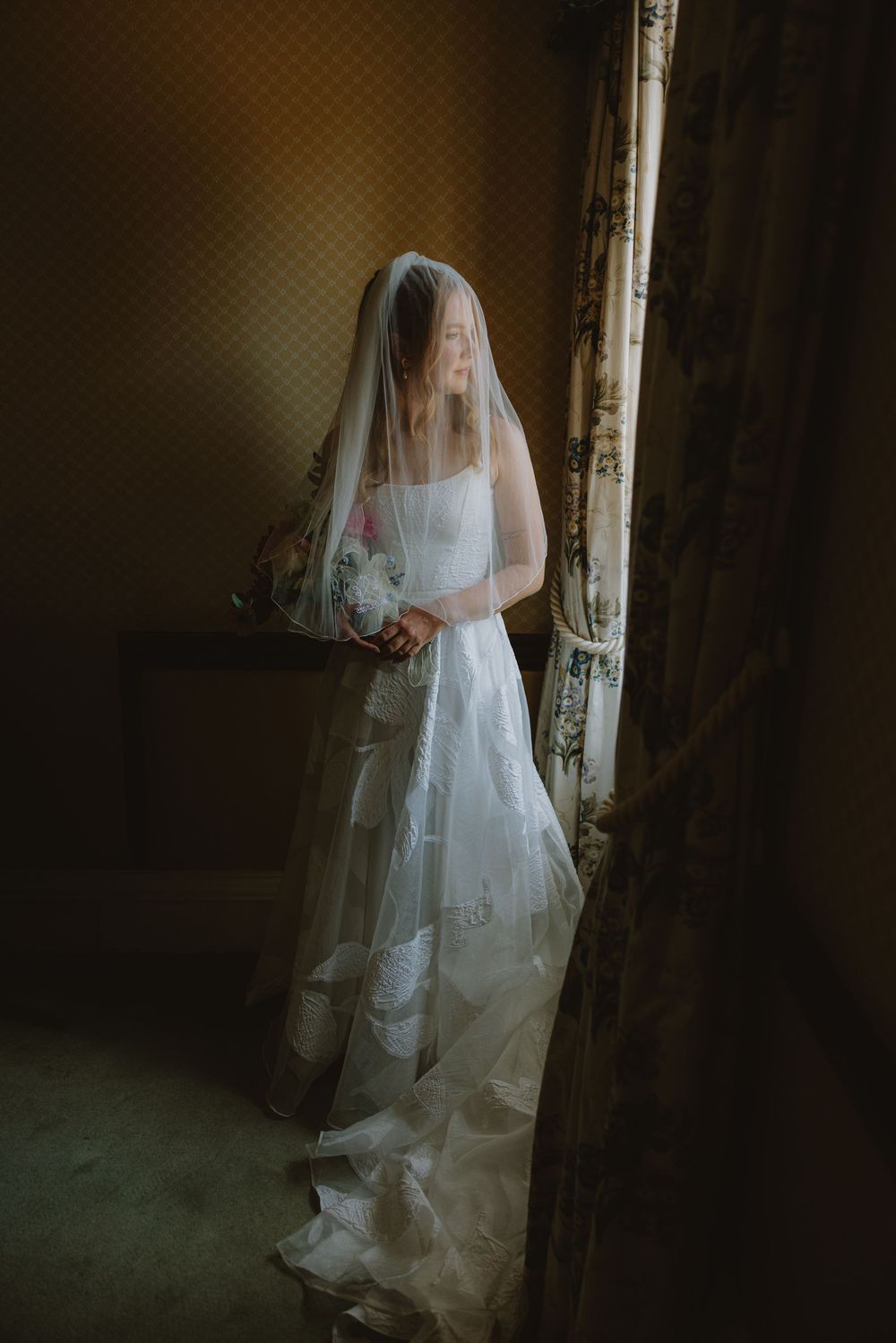 A bride in a white wedding dress and veil stands in dramatic lighting near a vintage curtained window.