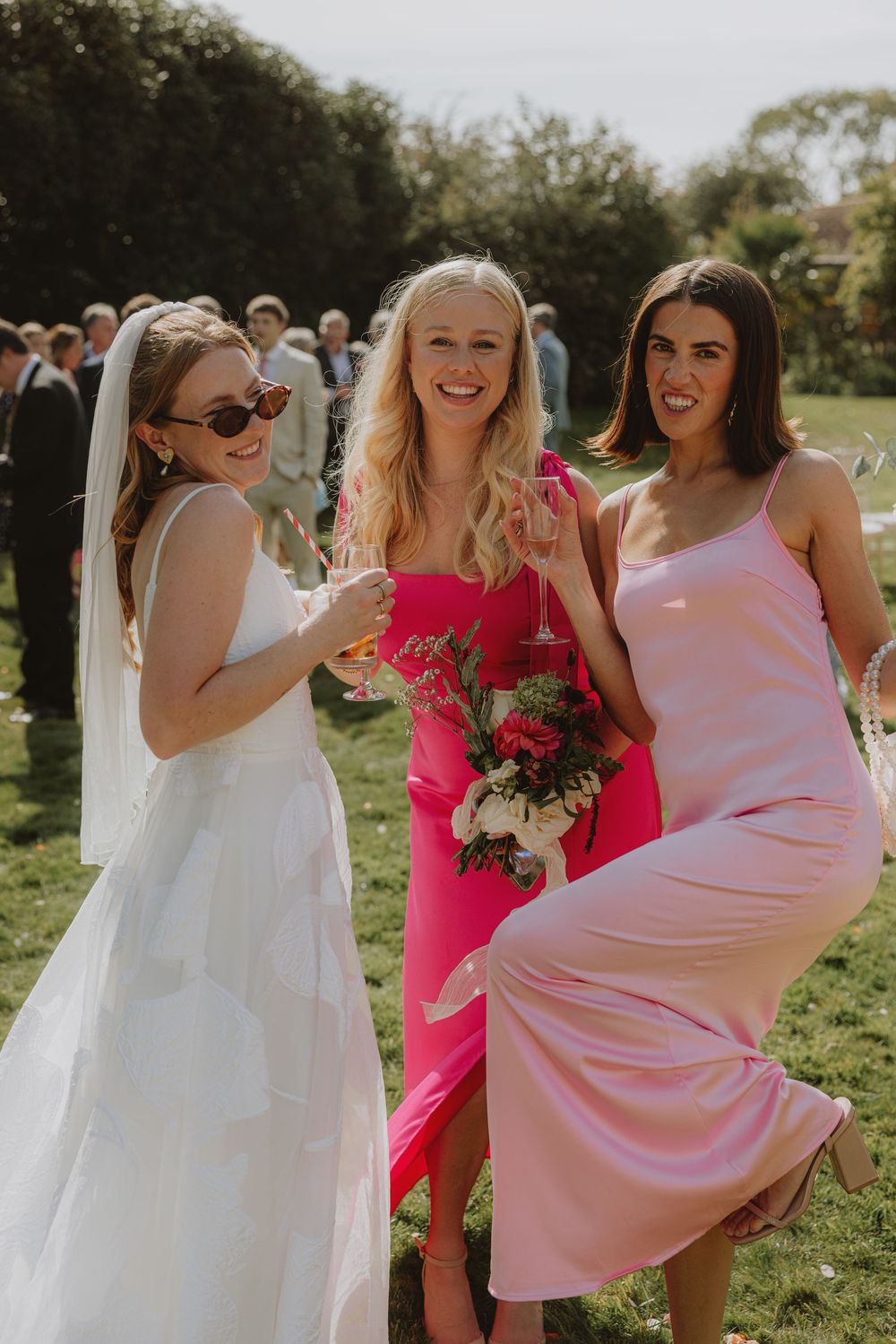 Three wedding attendees in pink and white dresses pose with flowers in a sunny garden.