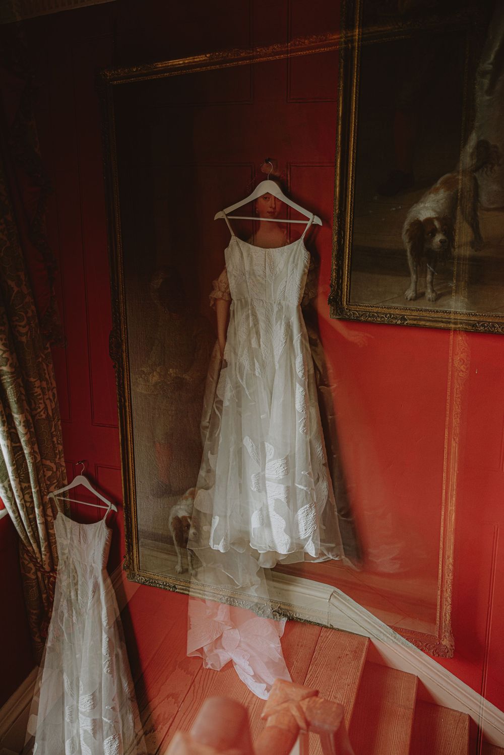 White wedding dress displayed on hangers against a red wall with ornate frames.