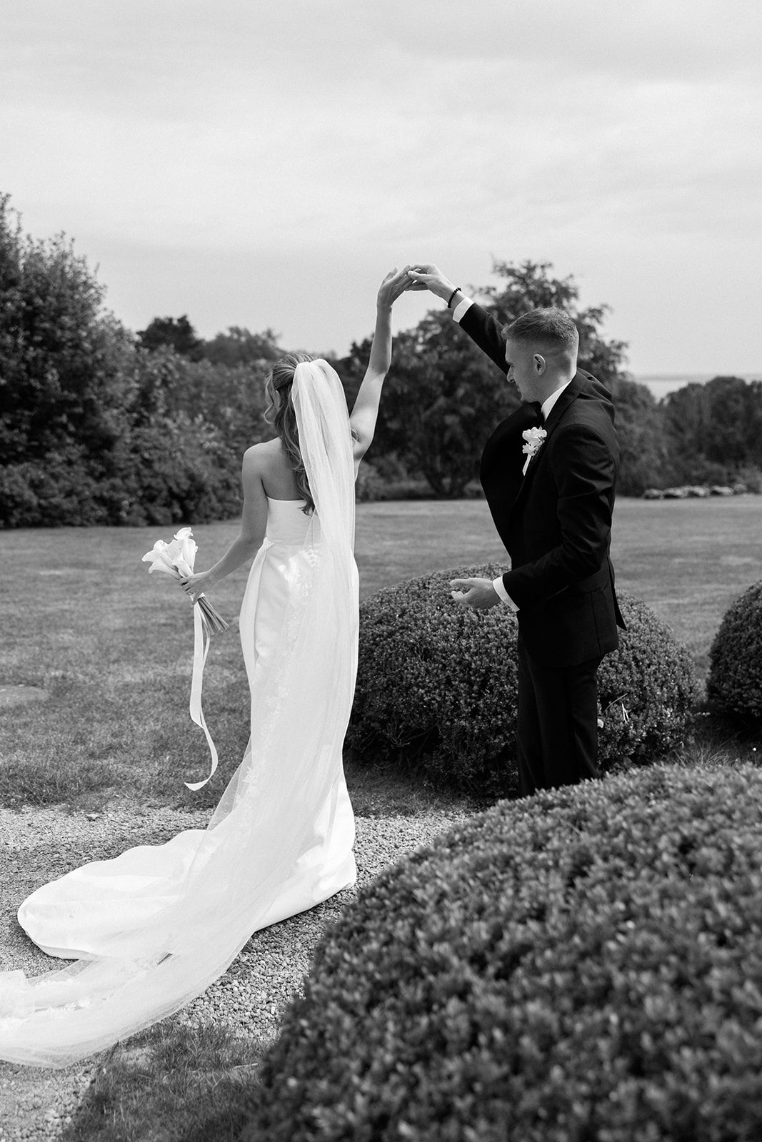 A black and white wedding photo showing a couple in formal attire standing outdoors among manicured hedges.