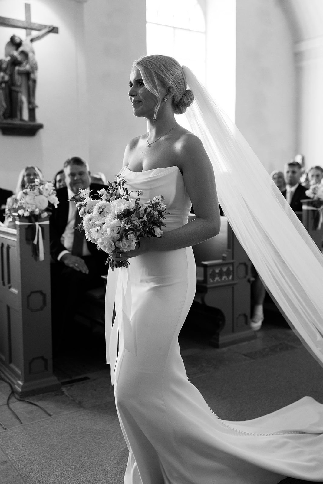 A bride in a sleek white wedding dress walks gracefully with her flowing veil trailing behind her in a church setting.