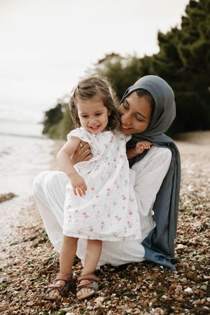 A tender moment captured on a beach covered in seashells and sand as a parent embraces a child in a white floral dress.