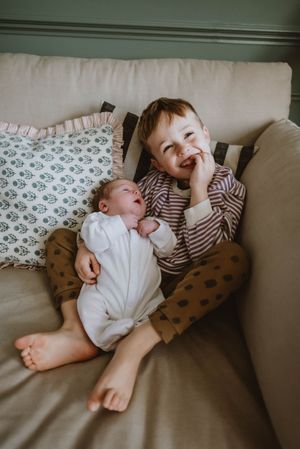 Two young children laugh and play together while lounging on a gray couch with patterned pillows.