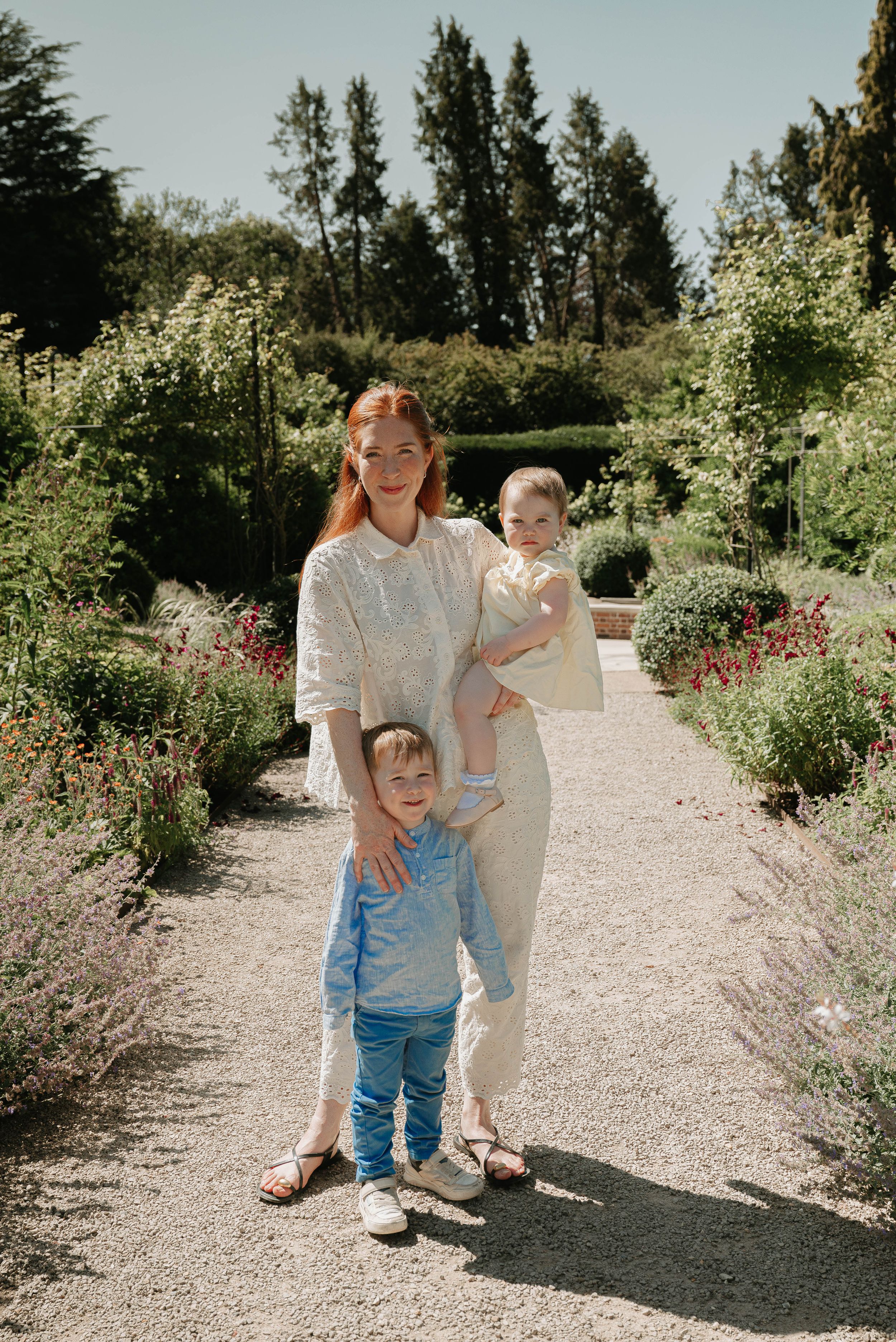 Family enjoying a sunny day walking along a garden path surrounded by blooming flowers and tall evergreen trees.