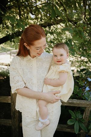 A tender moment captured outdoors as parent and baby share joyful expressions while sitting near a rustic wooden fence.