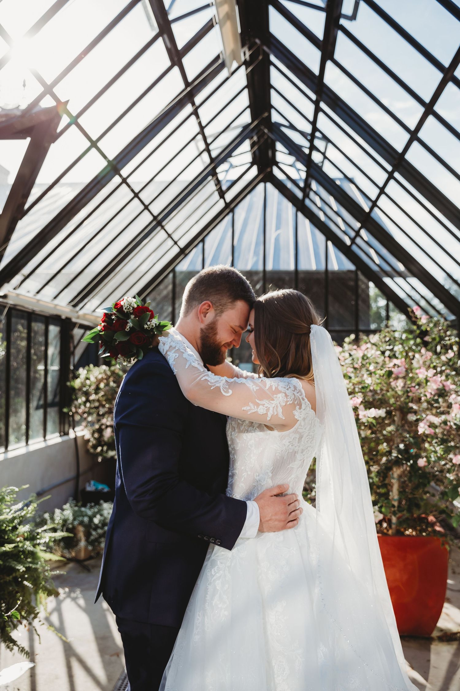 A couple shares an intimate moment in a sunlit greenhouse surrounded by blooming flowers and greenery.