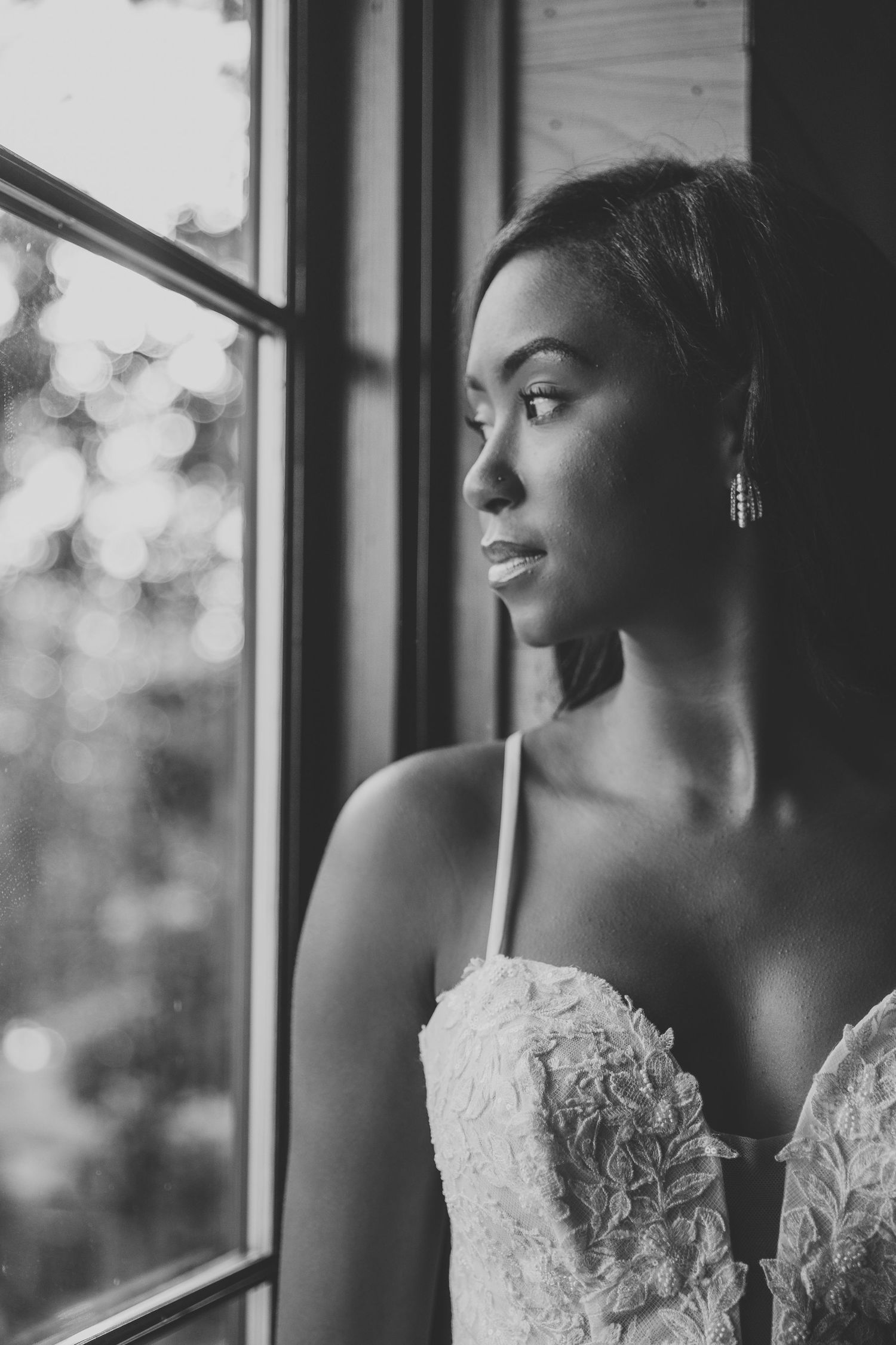 A bride in a lace wedding dress gazes thoughtfully through a window in a black and white photograph.