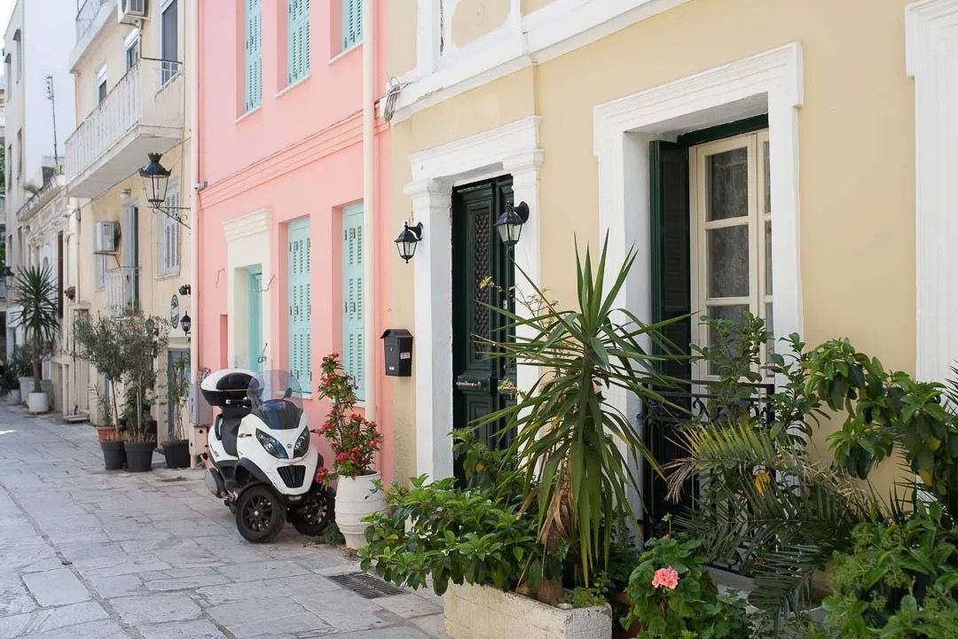Charming pastel-coloured buildings with potted plants line a narrow cobblestone street in a Mediterranean coastal town.