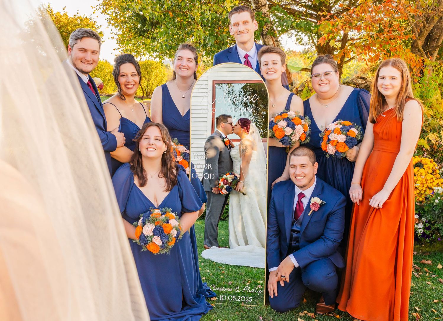A wedding party poses outdoors in front of vibrant autumn foliage with orange and yellow leaves in the background.