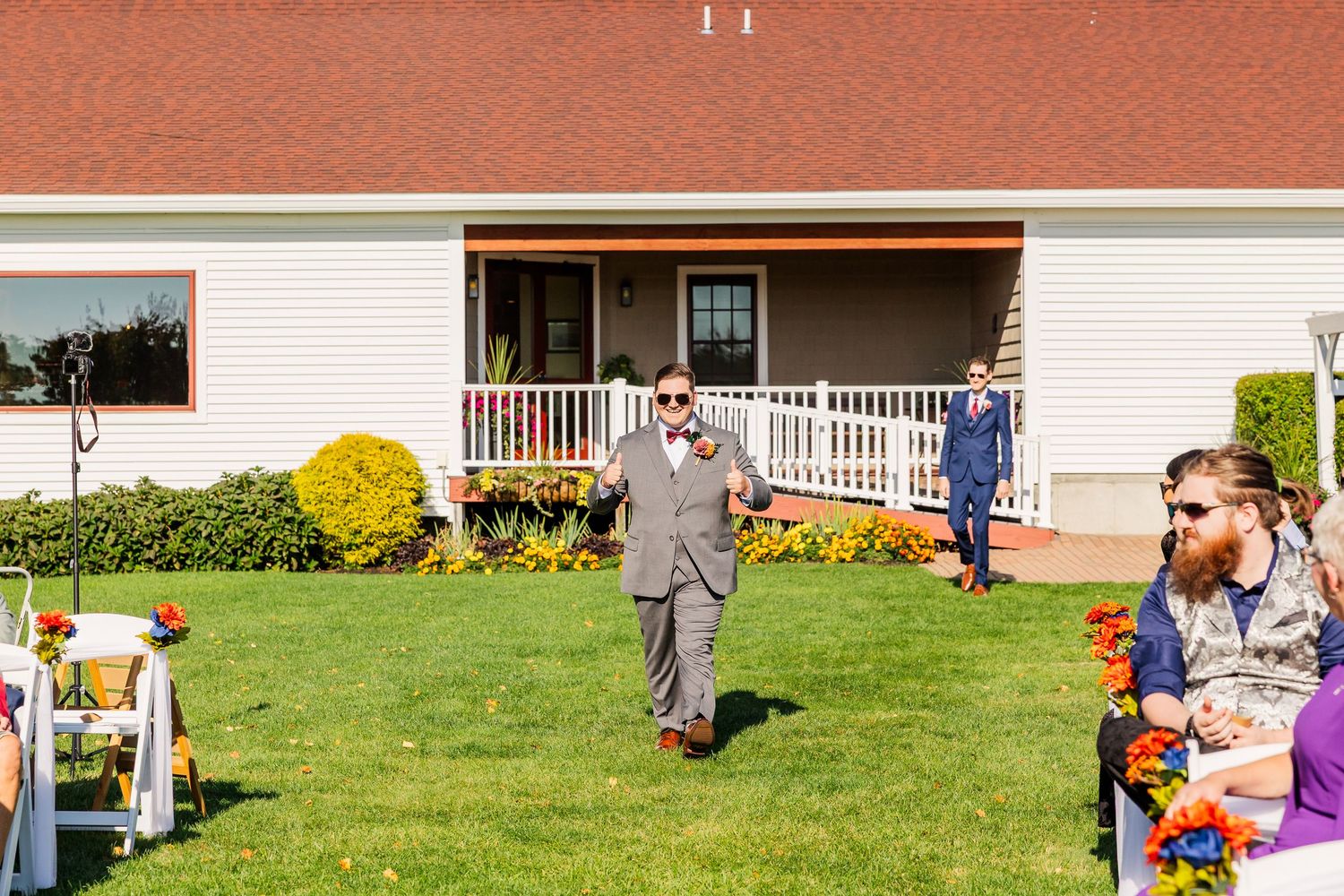 People gather outdoors at a backyard event with white chairs and a porch-style building in the background.
