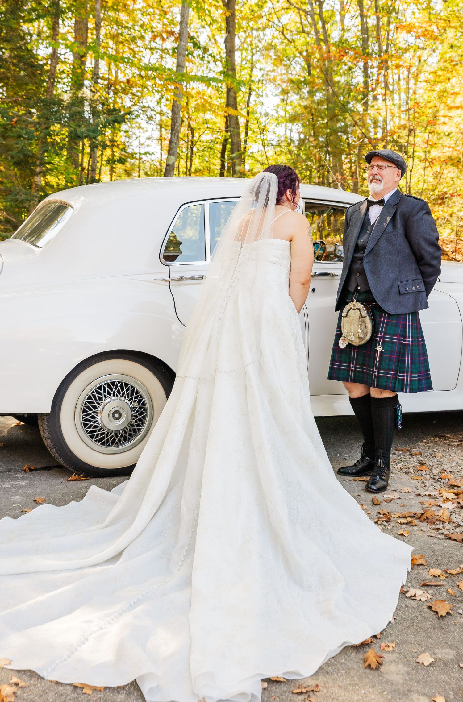 A couple poses near a classic white vintage car surrounded by autumn leaves and golden fall foliage.