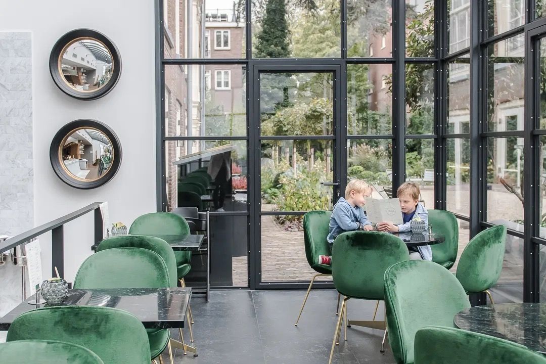 Modern café lounge with green velvet chairs and large windows overlooking a leafy courtyard
