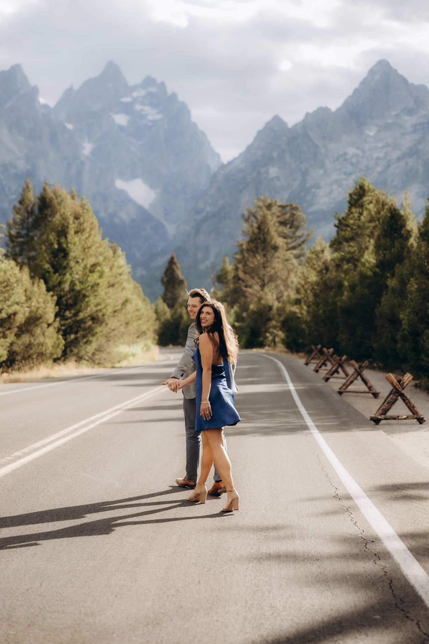 A couple walks down a mountain road with snow-capped peaks in the background during their winter wedding photoshoot.