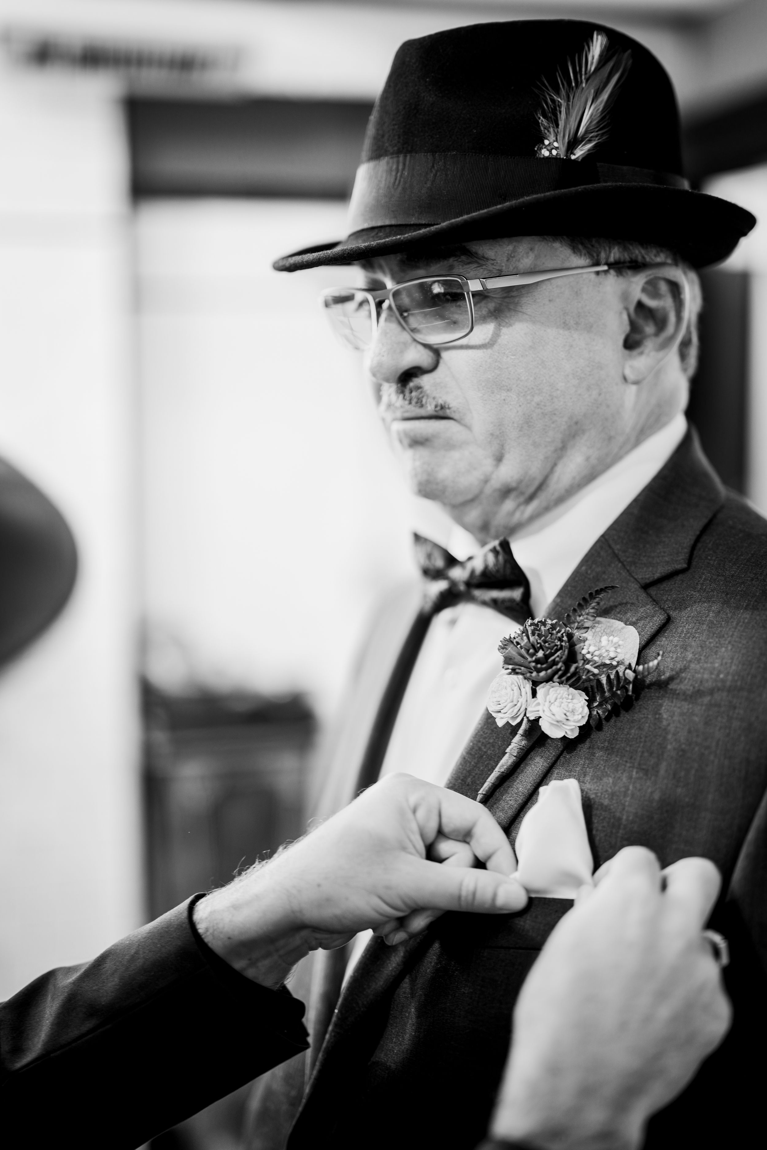 A formal black and white portrait of a gentleman wearing a fedora, bow tie and boutonniere being adjusted.