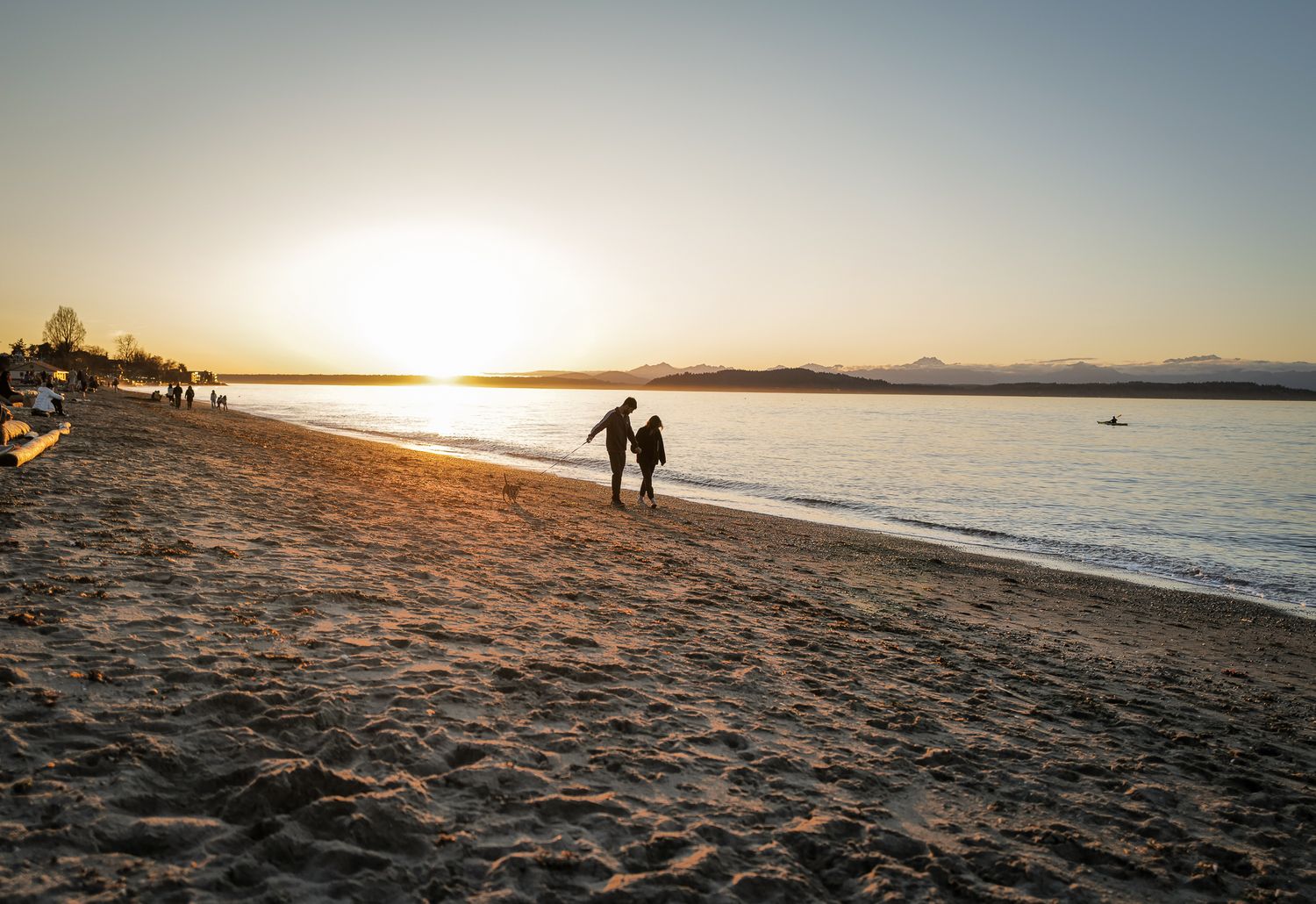 Couple walking hand in hand along a sandy beach at golden sunset with mountains in the distance.
