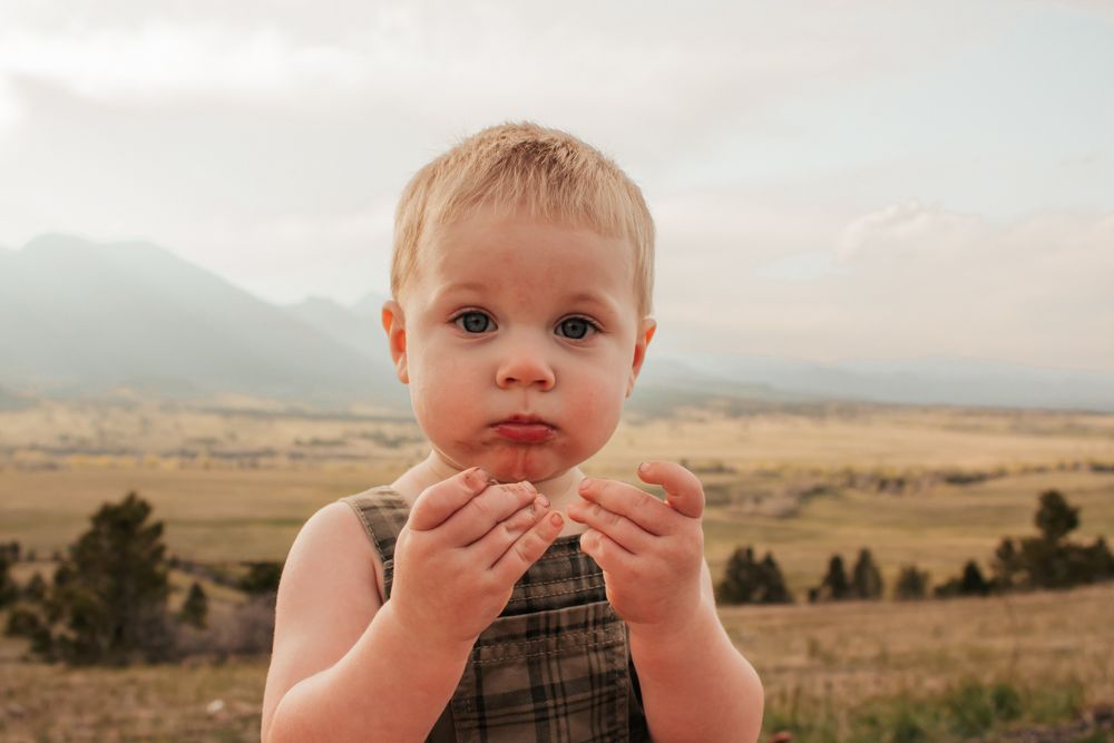 A laughing toddler in a plaid shirt stands outdoors against a scenic mountain landscape at sunset | Westminster Family Photographer