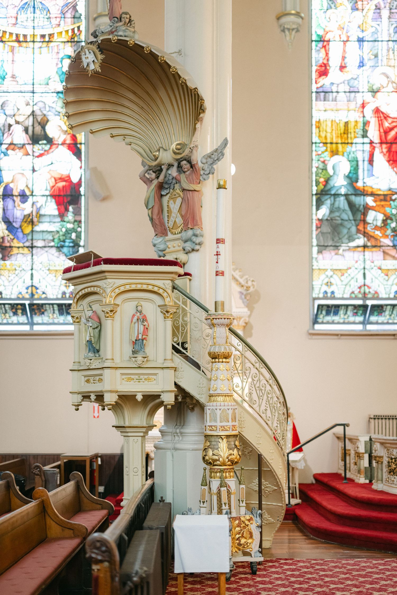 Ornate church pulpit with shell-shaped canopy and decorative staircase next to stained glass windows and red carpet steps.