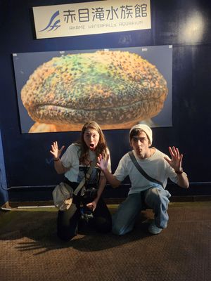 Two people pose playfully in front of an aquarium display sign with Japanese text at night.