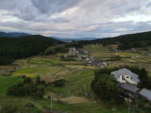 Scenic landscape of terraced rice fields and scattered houses in a mountainous Japanese countryside under cloudy skies.