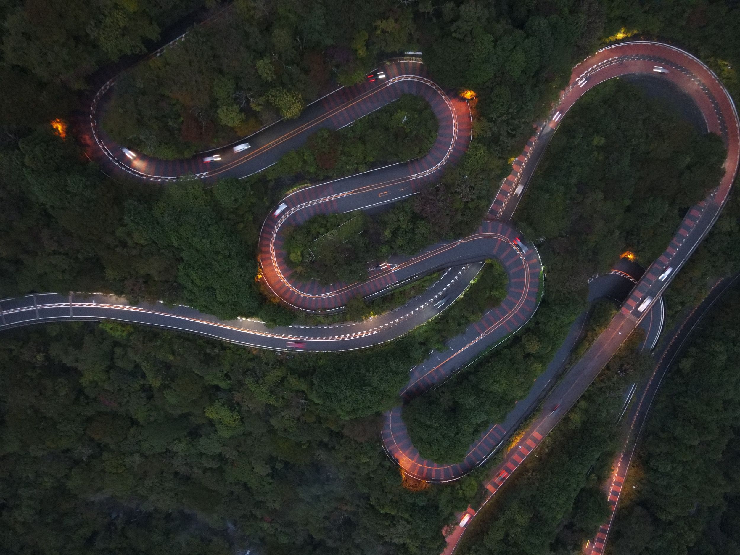 Aerial view of a winding mountain road with hairpin turns through dense forest at dusk.