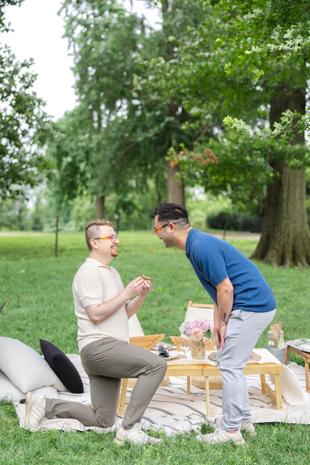 Two people enjoying a romantic outdoor picnic on a blanket in a lush green park setting.