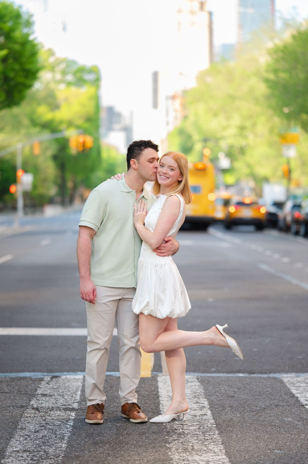 A romantic couple shares a tender moment on a tree-lined city street during a sunny afternoon.