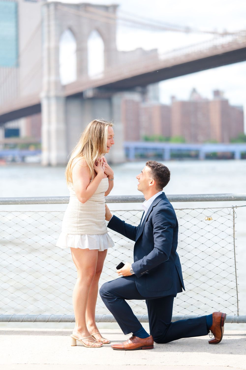 A romantic marriage proposal scene by the waterfront with the Brooklyn Bridge visible in the background.
