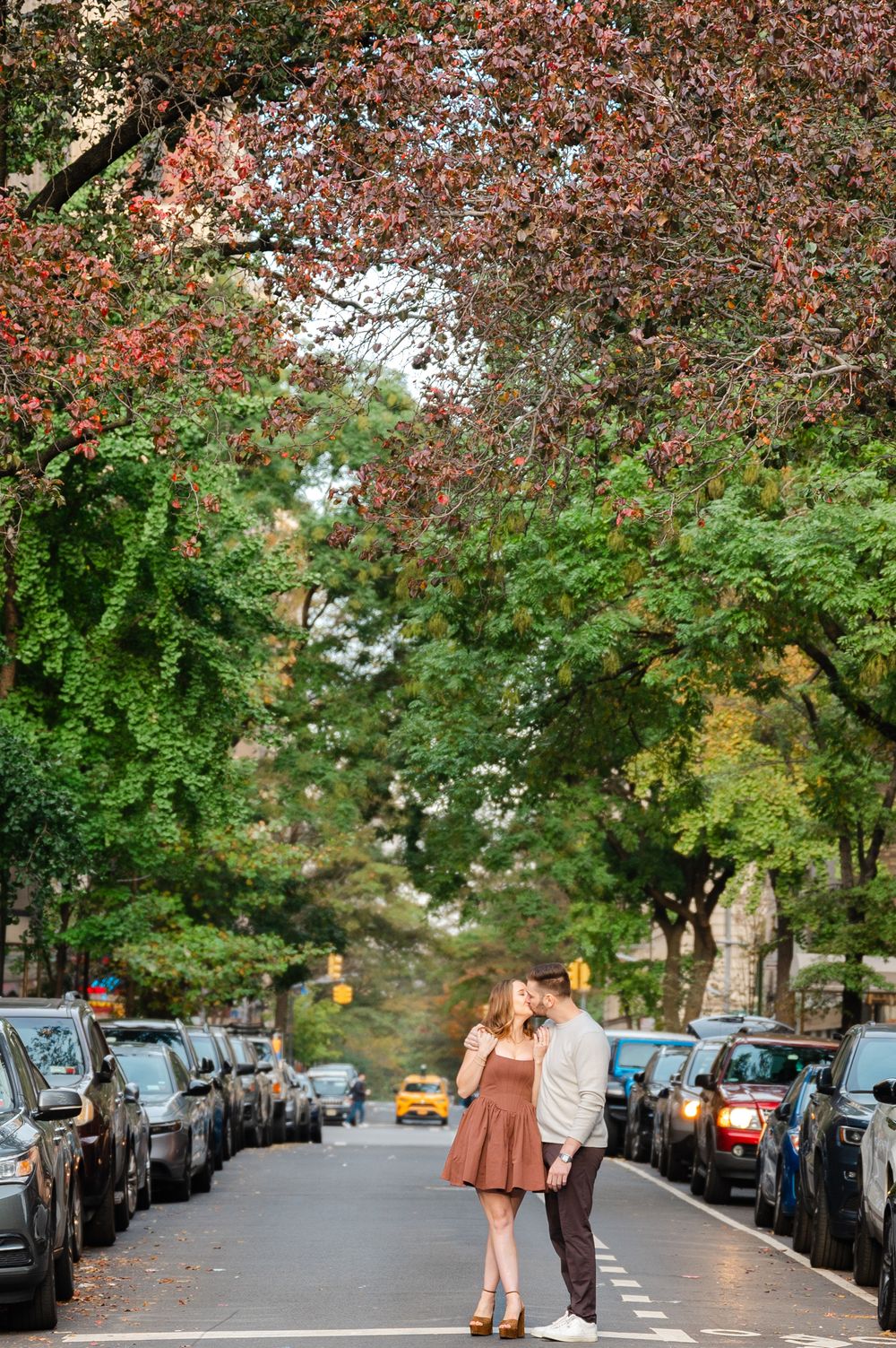 A couple stands in the middle of a tree-lined street with parked cars on both sides during autumn in New York City.