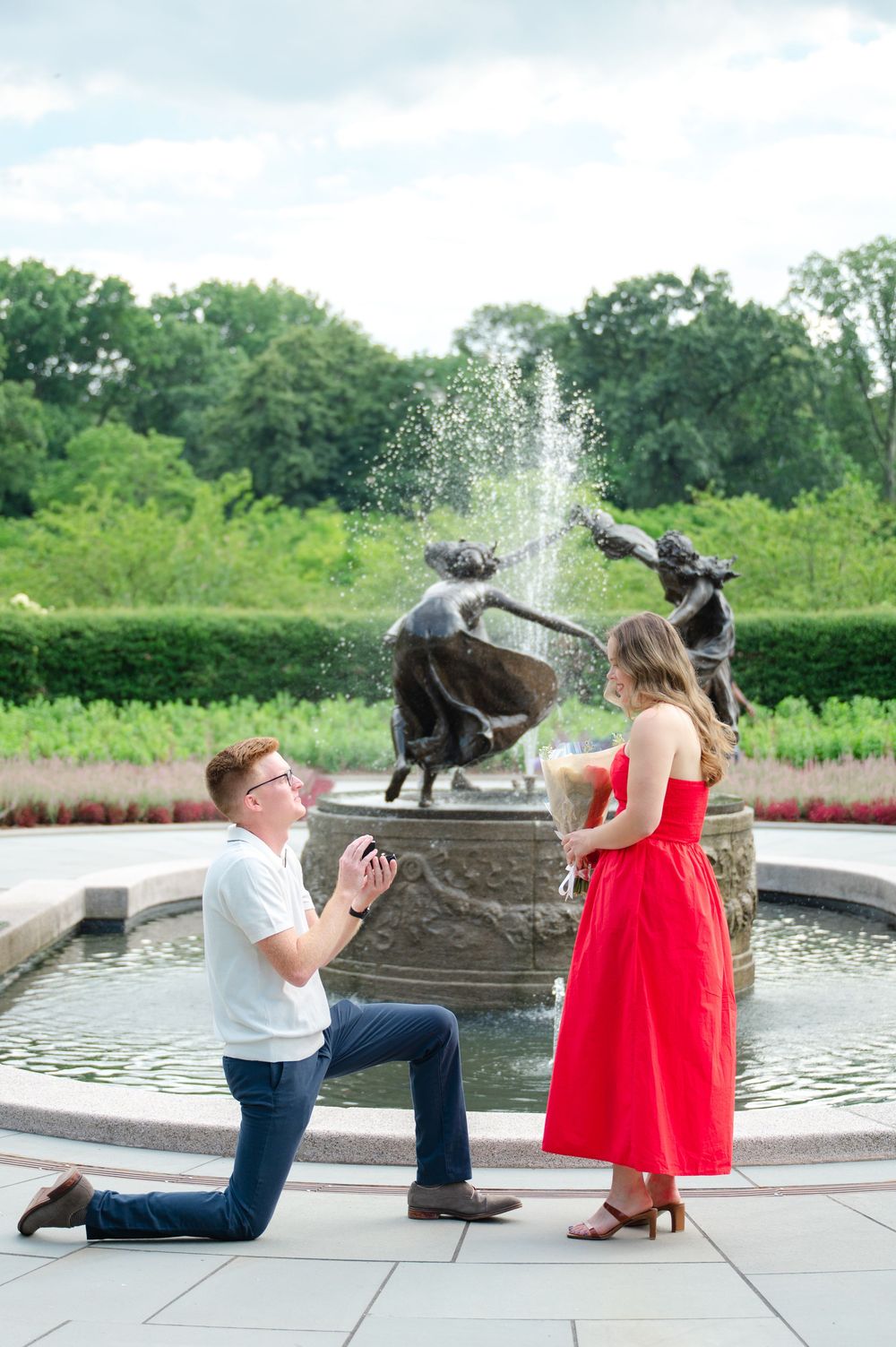 A romantic proposal scene by a fountain in a garden with decorative bronze sculptures on a sunny summer day.