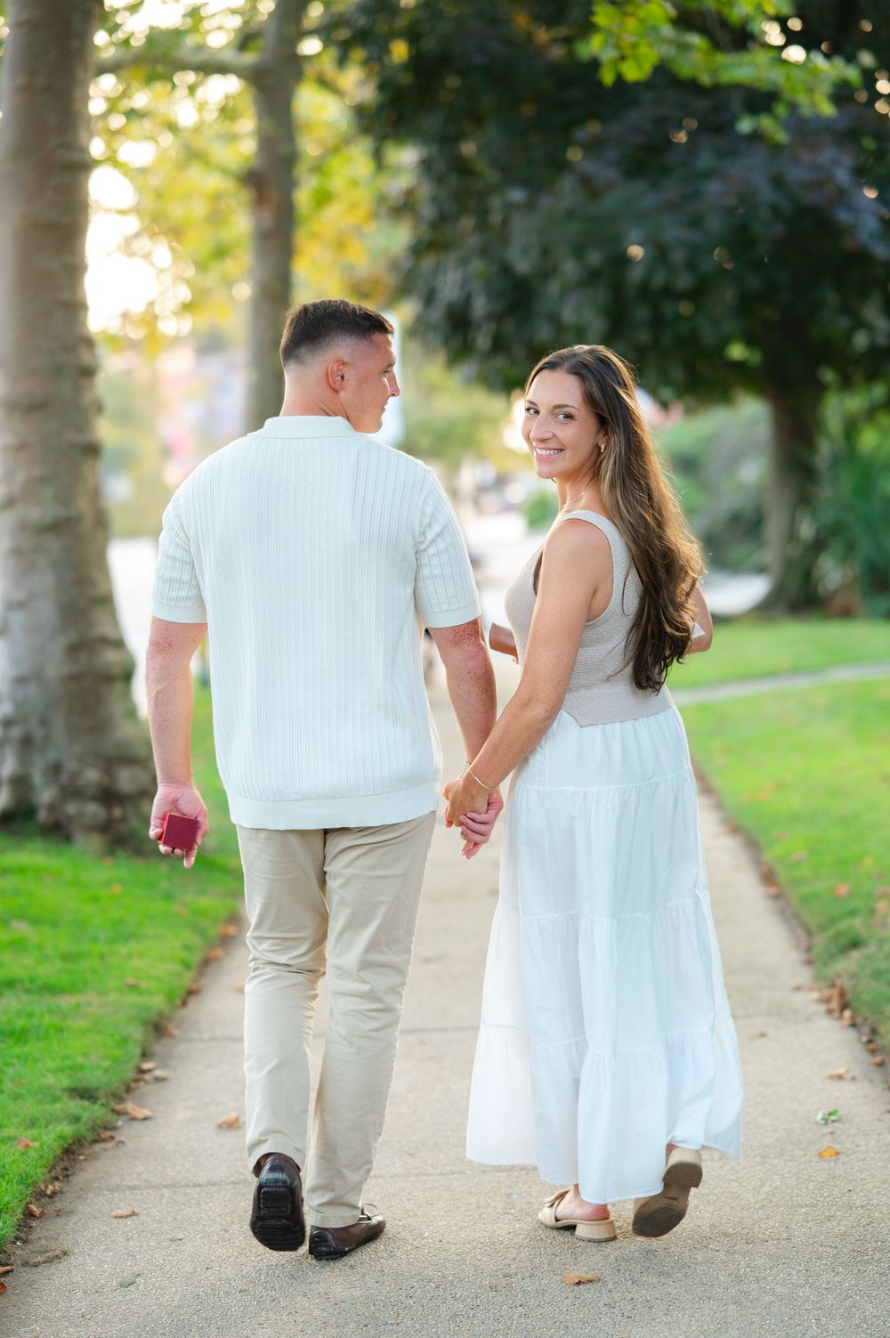 A couple walks hand in hand down a tree-lined path in summer, dressed in white and beige outfits.