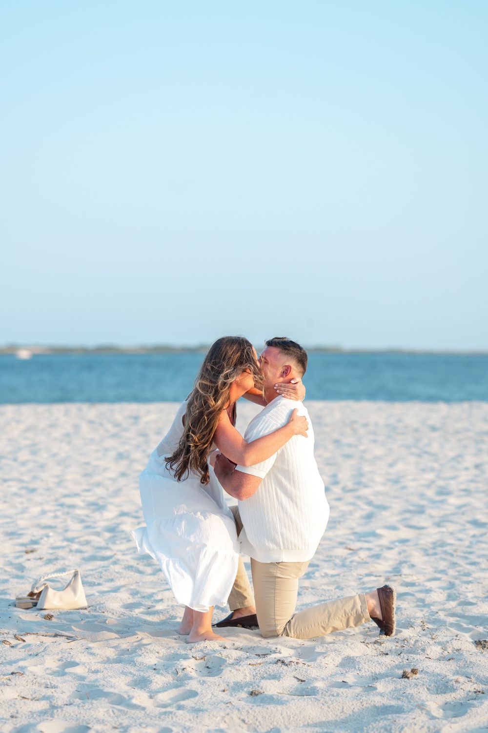 A couple shares a romantic moment while kneeling together on a pristine white sandy beach at sunset.
