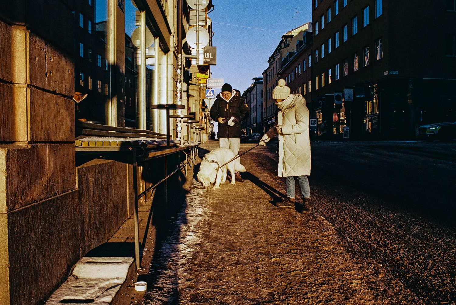 People stand on a snowy city sidewalk in winter sunlight along an urban street lined with buildings.
