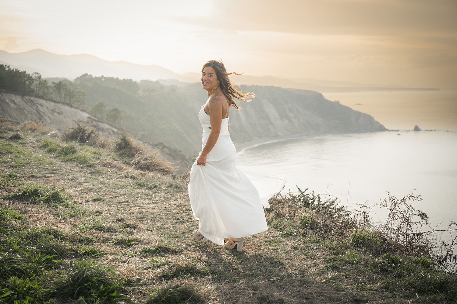 A flowing white wedding dress billows in the wind on a coastal cliff during golden hour sunset.