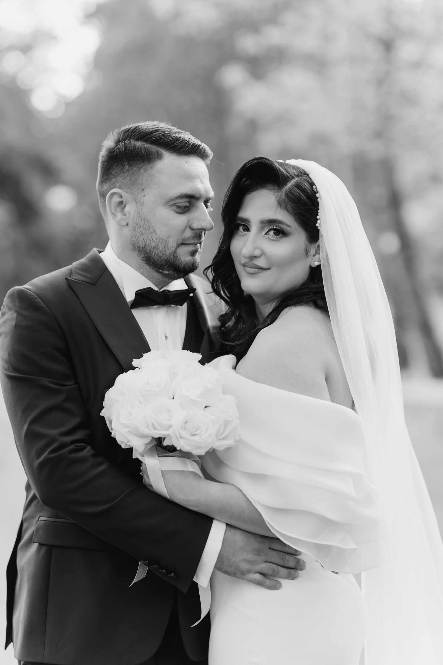 A bride in a white dress and groom in a black tuxedo walk through a sunlit forest during their wedding photoshoot.