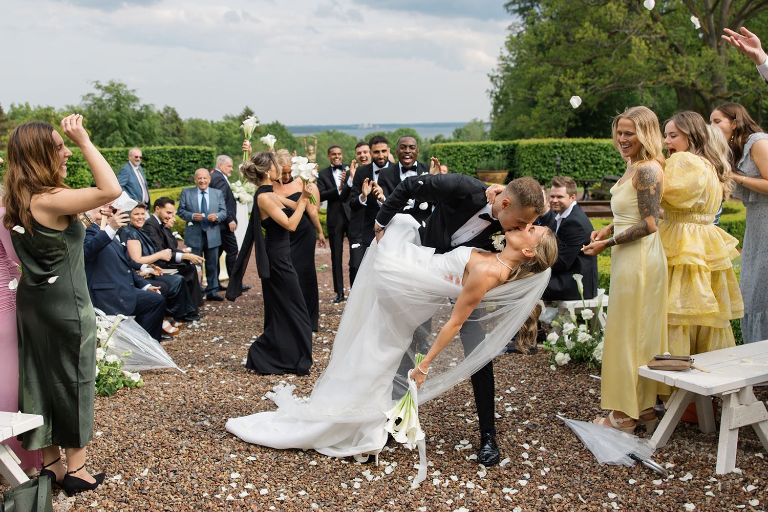 Wedding guests toss white paper streamers in celebration during an outdoor ceremony on a sunny day.