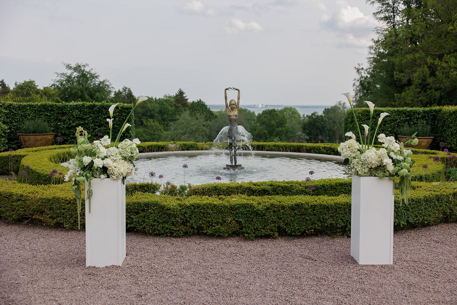 White floral arrangements in square planters line a curved garden path surrounded by manicured hedges and greenery.